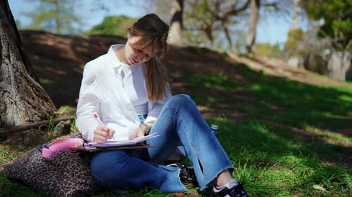 Konzentrierter Student, der Notizen schreibt, während er bequem im Gras im Schatten eines Baumes sitzt