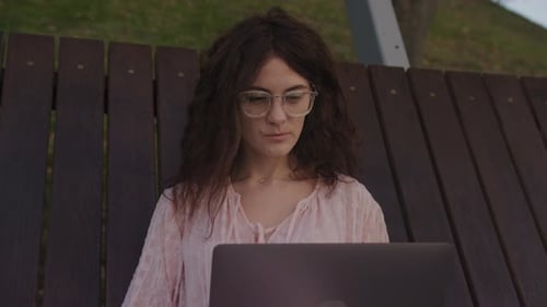 Handheld Close Up of a Curly Caucasian Woman Working on Her Laptop Computer on a Park Bench