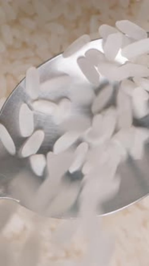 Vertical video. White rice being poured onto a spoon from above, a rotating background of grains.