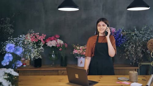 Woman Using Laptop and Phone in Flower Shop Surrounded By Plants and Flowers A Call From a Seller to