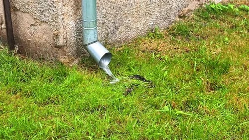 Rainwater flowing from metal downspout onto green grass next to old building wall