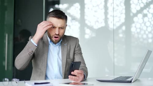 Frustrated businessman reads bad news on a smartphone while sitting at workplace in business office.