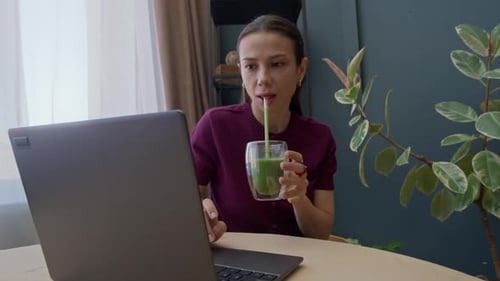 Woman Drinks Green Smoothie While Using Laptop
