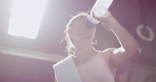 Active young woman drinking water during a break from exercise in a gym from below