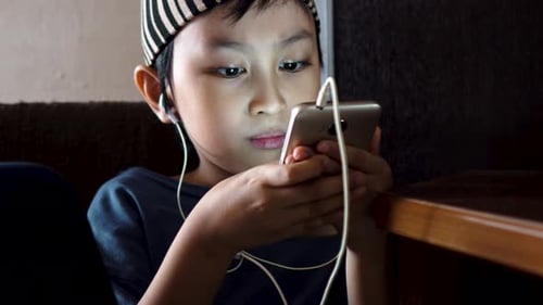 Young Southeast Asian Muslim boy watching videos on Handphone, alone indoors in dimly lit room.