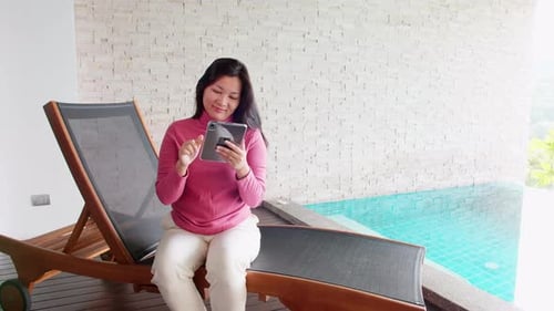 Asian woman relaxing on a pool bed and using digital tablet near the private pool during her morning