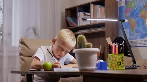 A Schoolboy is Sitting in a Chair at a School Desk and Writing in a Notebook