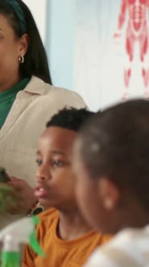 Vertical video: Entering teacher in green top holding tablet, teaching boy in classroom with chart