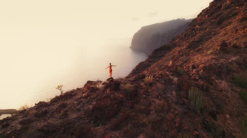 Woman in orange activewear stands on a rocky cliff edge, admiring the sunset over the ocean in Tener