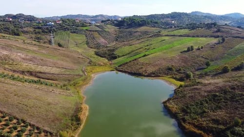 Aerial above beautiful small lake inside the albanian countryside with agricultural fields.