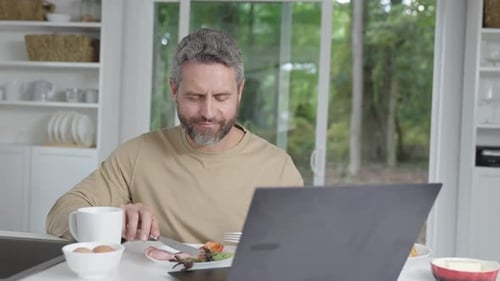 Man Eating Breakfast and Using Laptop in Kitchen