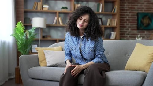 Woman Massaging Knee in Living Room, Experiencing Pain