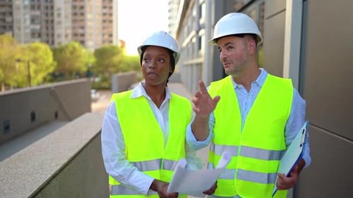 Two diverse architects with helmets and safety vests reviewing project plans outside office