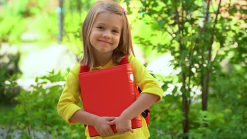 Portrait of Little Student Girl Wih Book Looks at Camera and Laughs in Schoolyard