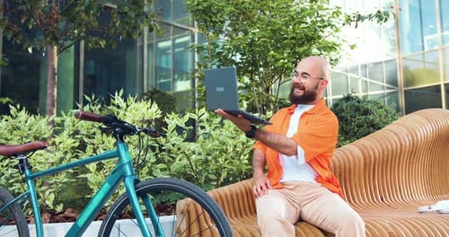 Moderno hombre urbano milenario en una videollamada en línea con una computadora portátil, sentado al aire libre junto a la bicicleta.