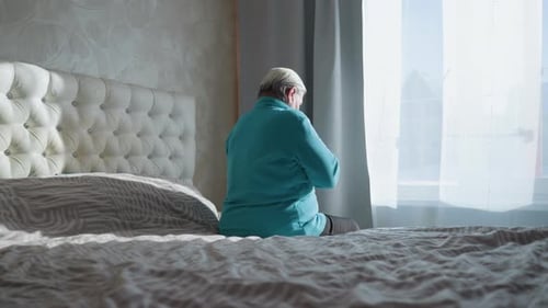 Old Woman in Green Sitting on Bed Reflecting By Window in Soft Sunlight