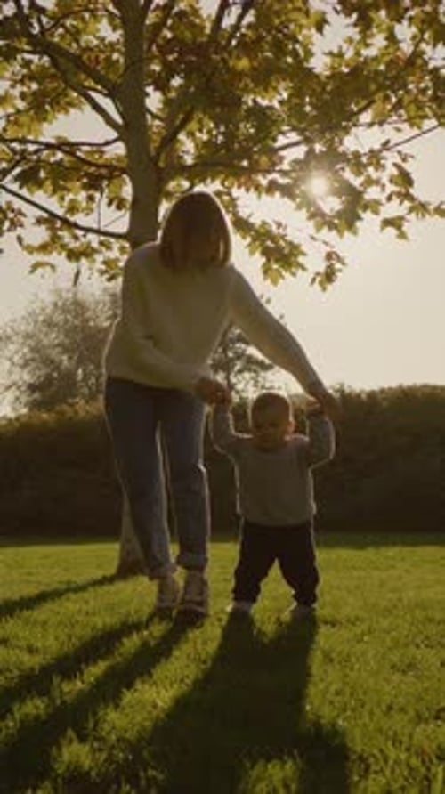 Vertical video Mother guides baby boy taking first steps on a sunny park