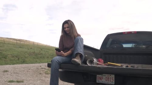Woman Uses Phone Sitting on Truck in Field