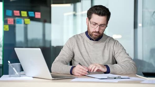Businessman reviewing document and signing contract while sitting at desk in modern office. Mature