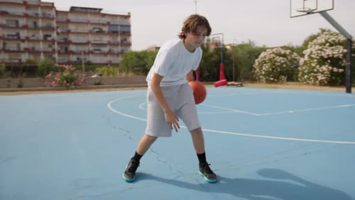 Young Blonde Athlete Dribbles Under His Legs In Outdoor Basketball Court