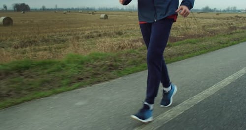 Close Up on Male Jogger Legs Running on a Road of a Countryside Field During Day. Motivated Mid