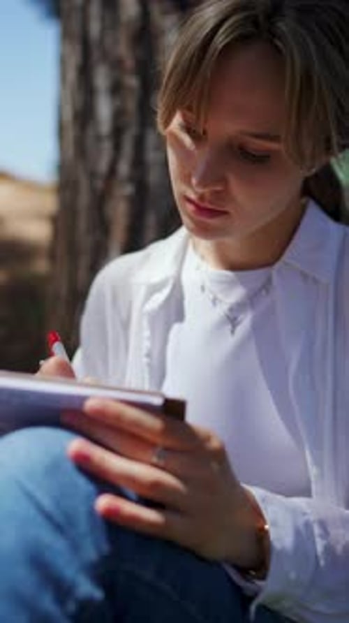 Female Student Scribbling Notes with Red Pen Leaning Against Pine Tree Trunk During Quiet Park Study