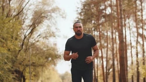 Young Muscular Jogger with a Focused Face Running on the Path of a City Park at a Steady Pace on a