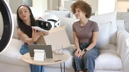 African American female hosts in living room unboxing white VR headset with left host reaching box