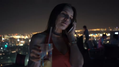 Young Woman Talking on Cellphone and Drinking Cocktail on Terrace in Bar