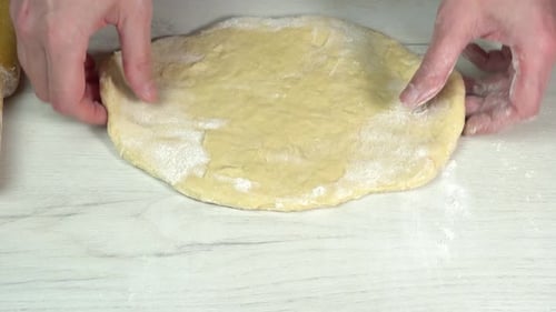 Hands turn rolled out dough and sprinkle it with flour. The cook uses a rolling pin to stretch