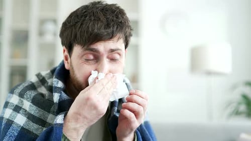 Sick man suffering from a cold wrapped in blanket sitting on sofa in living room at home.