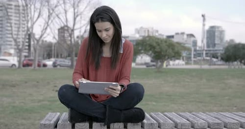 Woman with Ipad outdoors sitting on a bench in the park.