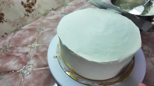 Eine Hand trägt eine Schicht Zuckerguss auf den Kuchen auf. Auf dem Tisch eine Schüssel Zuckerguss und ein Messer
.