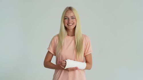 Studio Portrait of Cheerful Blonde Young Woman with Broken Arm Wrapped in White Plaster Bandage