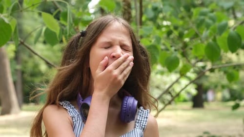 Yawning Tired Kid Girl with Headphones in City Park on Green Nature Background