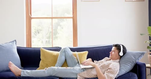 Happy biracial man wearing headphones lying on couch using laptop at home, copy space, slow motion
