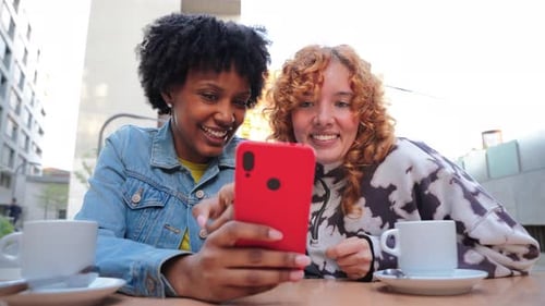Excited Young Women Sharing Joyful Moments While Looking at Smartphone Together at a Cafe Table