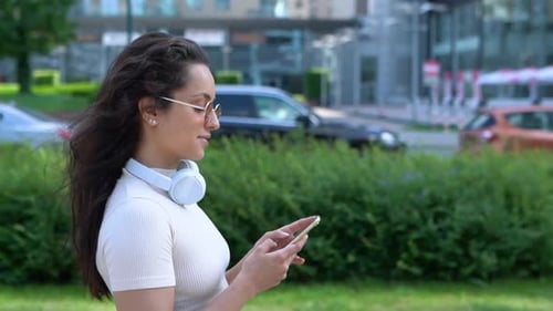 Close Up of Caucasian Pretty Cheerful Female with Headphones Walks in City and Smiling Texting on