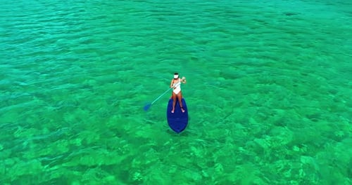 Luftaufnahme einer Frau in weißer Badesuite, die auf einem SUP-Board mit Paddel auf kristalltürkisfarbenem Wasser steht