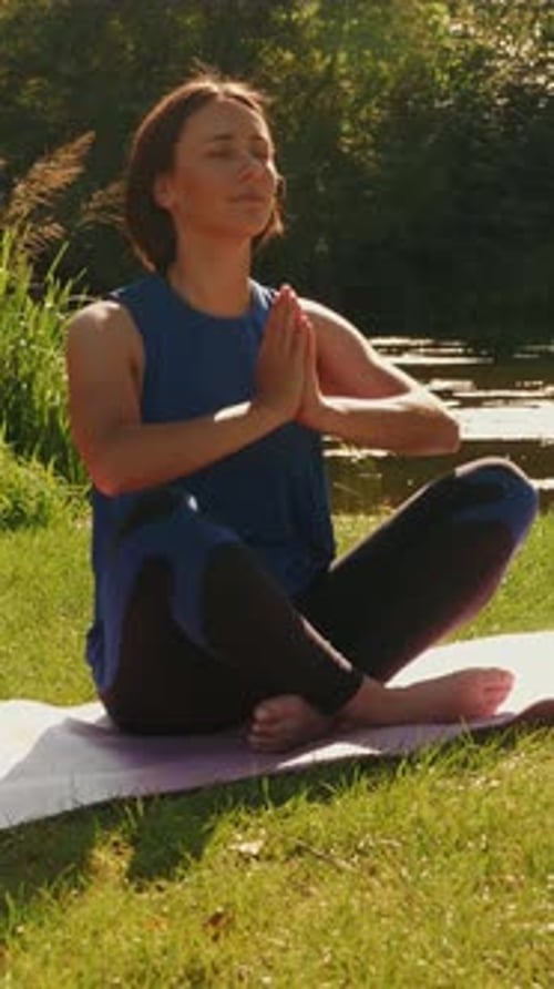 Woman sitting on yoga mat in lotus position at park near river. Vertical video