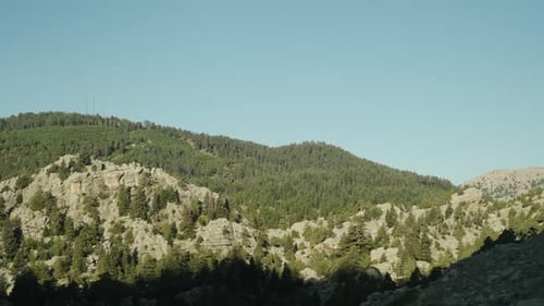 A view from a moving car of mountains and trees on rocky cliffs, under clear skies. Slow motion.