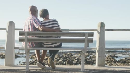 Happy senior african american couple sitting on bench on promenade by the sea, slow motion