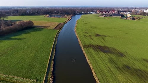 Luftaufnahme eines ruhigen Kanals, der zwischen Grasfeldern verläuft, mit Schilf entlang des Ufers, eingezäunter Weide