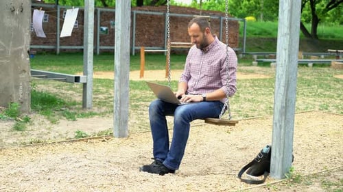 Young man working on laptop enjoying the fresh air in the park on a swing