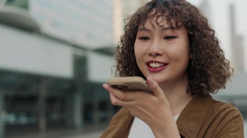 Happy Asian Woman With Curly Hair Recording Voice Message On Her Phone While Standing In Center