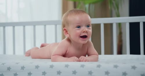 Baby Boy with Blue Eyes with Short Hair Lying on Stomach in Diaper in His white Crib and Smiling