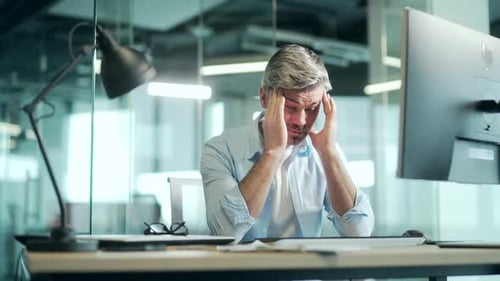 Exhausted, overworked mature office worker sitting in the workplace with a severe headache massaging