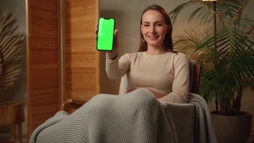 Happy Smiling Woman Shows a Green Mobile Phone Screen While Looking at a Camera on a Chair a