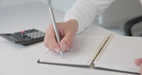 Nahaufnahme von Händen: Ein Mann in einem Hemd zeichnet ein Diagramm und macht sich Notizen mit einem Stift in einem Notizblock auf einem Metall