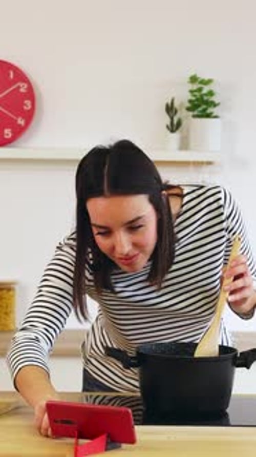 Vídeo de una joven feliz cocinando y siguiendo la receta en el teléfono en la cocina